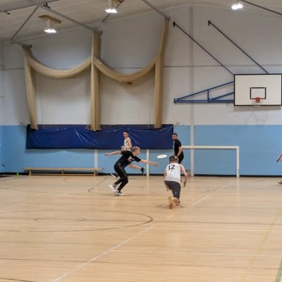 A disc mid-flight indoors as two players close in to receive it near the sports hall's basketball hoop