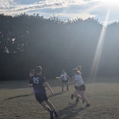 A player throwing a yellow disc past the goalposts on a sunny outdoor pitch, teammates close by